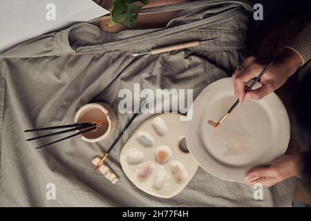 De dessus crop femelle artisan peinture ornement sur vaisselle en céramique sur table avec tissu et outils dans studio créatif Banque D'Images