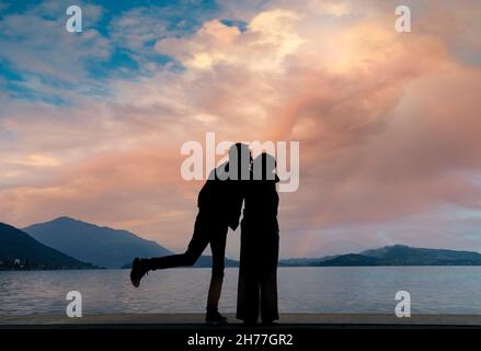 Silhouette d'un couple mignon amoureux au moment de l'embrasser avec un fond d'un lac au coucher du soleil avec des nuages de tons chauds illuminés par le soleil. Banque D'Images