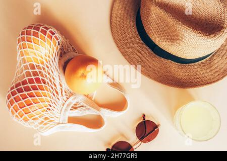 Oranges dans un sac à cordes, verre avec limonade, chapeau de plage et lunettes de soleil sur fond clair, vue de dessus, plat. Banque D'Images