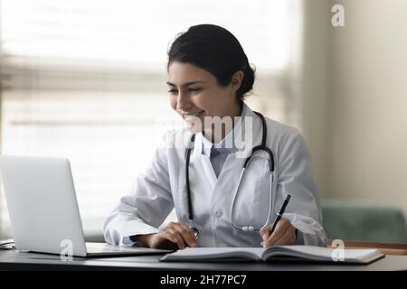 Femme indienne souriante médecin médecin utilisant un ordinateur portable, prenant des notes Banque D'Images