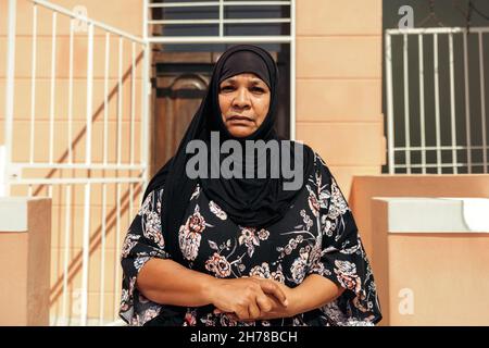 Femme musulmane d'âge moyen regardant la caméra tout en se tenant devant sa maison pendant la journée.Une femme d'âge moyen confiante porte un hijab et Banque D'Images