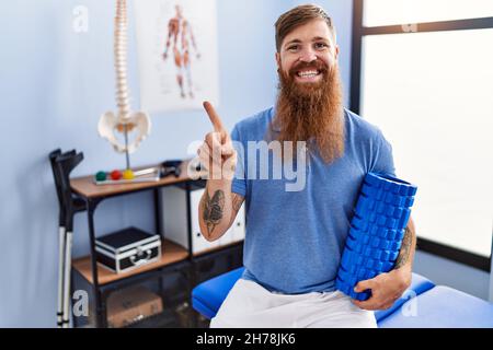 REDHEAD homme avec la longue barbe tenant le rouleau en mousse à la clinique médicale souriant heureux de pointer avec la main et le doigt sur le côté Banque D'Images