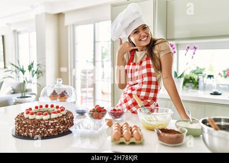 Belle jeune brunette pâtissière chef femme cuisant des pâtisseries à la cuisine souriant faisant un geste de téléphone avec la main et les doigts comme parler sur le télép Banque D'Images