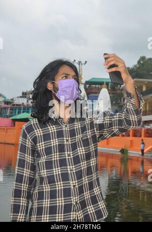 Un jeune homme d'asie du Sud avec de longs cheveux prenant le selfie ou ayant un appel vidéo avec son smartphone avec le port du masque facial en se tenant debout à l'extérieur Banque D'Images