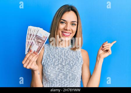 Jeune femme blonde caucasienne portant des billets en livres égyptiens souriant, souriante, pointant avec la main et le doigt sur le côté Banque D'Images