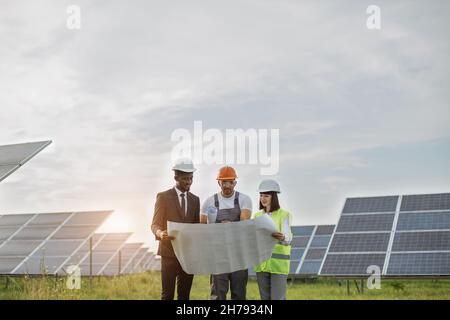 Groupe de personnes multiethniques et de casques de sécurité regardant la ferme solaire et regardant les dessins.Deux ingénieurs et un technicien examinant le plan de construction photovoltaïque à l'extérieur. Banque D'Images