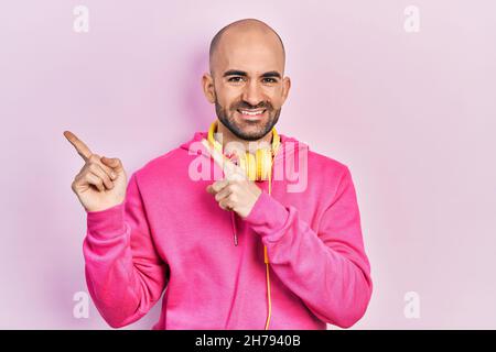 Jeune homme chauve portant des vêtements de gym et utilisant des écouteurs souriant et regardant la caméra pointant avec deux mains et des doigts sur le côté. Banque D'Images