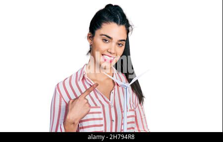 Jeune femme hispanique tenant un moulin à vent solaire pour l'électricité renouvelable souriant se réjouit de pointer du doigt et de la main Banque D'Images