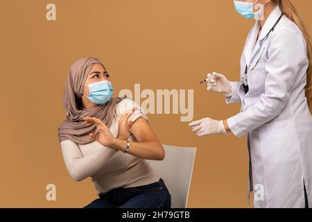 Jeune femme arabe dans le masque de visage peur de faire l'injection de vaccin de coronavirus, faisant UN GESTE D'ARRÊT et regardant le médecin Banque D'Images