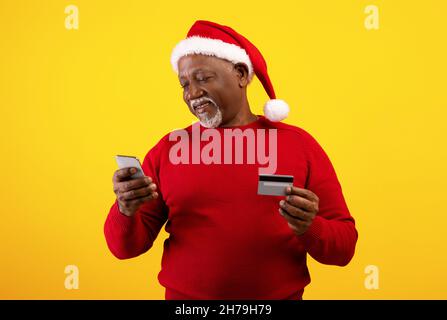 Joyeux homme noir senior avec téléphone portable et carte de crédit portant chapeau de Santa, acheter des cadeaux de Noël en ligne Banque D'Images