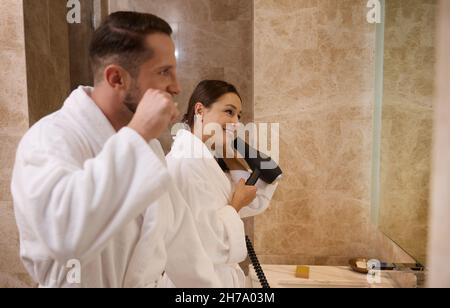 Portrait latéral de beau jeune couple hétérosexuel amoureux, portant des peignoirs blancs en éponge se brossant les dents et séchant les cheveux debout devant un bain Banque D'Images