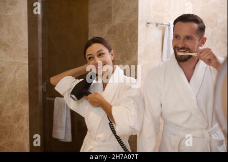 Réflexion dans la salle de bains miroir de jeunes heureux hétérosexuels couple dans l'amour pendant l'hygiène du matin.Un homme charmant se brossant les dents tout en se tenant à côté du salut Banque D'Images