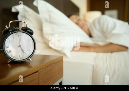 Mise au point douce sur le réveil noir sur la table de nuit à côté d'un lit flou avec une femme endormi.Espace de copie pour la publicité.Sommeil sain et réveil conce Banque D'Images