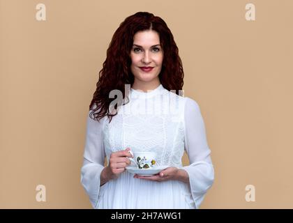 Femme avec le sourire doux tient Teacup isolé contre beige arrière-plan belle femme souriante en robe blanche romantique tient le teacup vintage. Banque D'Images