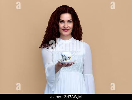 Femme offrant une tasse de thé avec sourire doux isolé sur fond beige.Portrait en gros plan d'une belle femme souriante en robe blanche tenant une tasse à thé. Banque D'Images