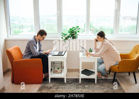 Vue latérale d'un couple travaillant de chez lui.Femme parlant au téléphone tout en enregistrant des données sur ordinateur portable, homme prenant des notes.Ils sont tous deux assis sur des fauteuils Banque D'Images