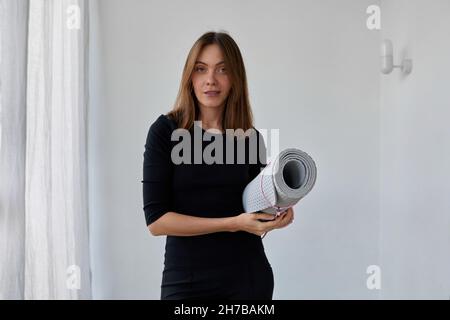 Une belle fille dans des vêtements noirs se tient dans la pièce blanche et tient un tapis de yoga dans ses mains tout en se tenant debout Banque D'Images