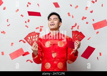 Joyeux homme asiatique en costume oriental traditionnel tenant des enveloppes rouges ou Ang Pao en fond gris avec des confetti, les textes chinois signifient grand Banque D'Images