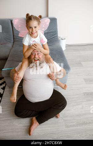 Une fille aux ailes roses est assise sur les épaules d'un père plumped au sol dans la salle de séjour Banque D'Images