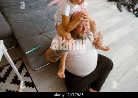 La fille est assise sur les épaules d'un père positif dans la salle de séjour Banque D'Images