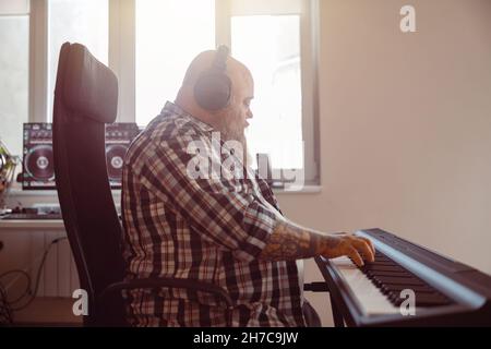 Homme de grande taille avec barbe et casque compose de la musique sur synthétiseur dans un studio bien équipé Banque D'Images