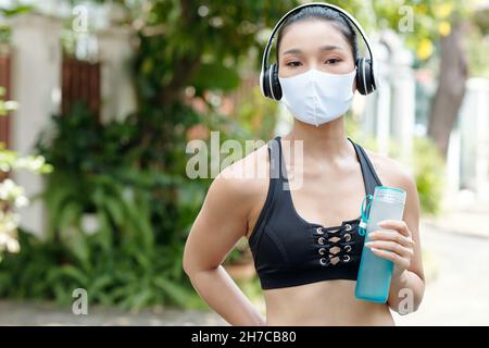 Jeune sportswoman dans un masque médical tenant une bouteille d'eau douce à boire après l'entraînement à l'extérieur Banque D'Images