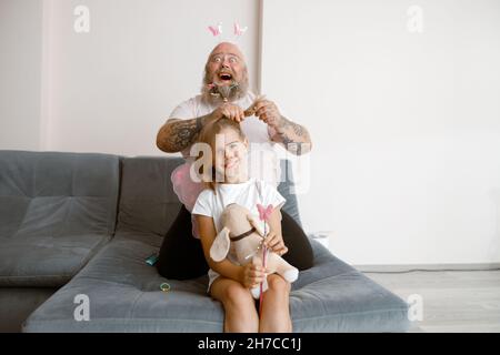 Plump Daddy fait de la queue de cheval à sa fille avec des ailes de fées et un chien jouet dans le salon Banque D'Images