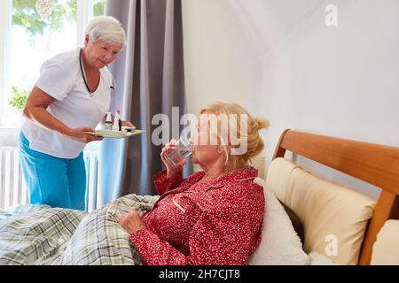 Une femme du service de soins infirmiers distribue des médicaments à une femme âgée malade au lit à la maison ou à la maison de retraite Banque D'Images