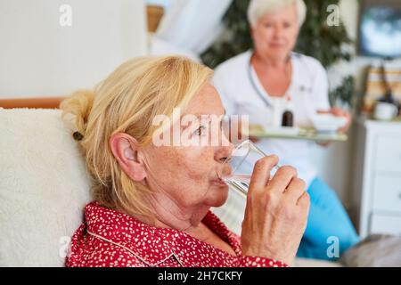 Une femme âgée malade prend des médicaments avec un verre d'eau dans les soins à domicile Banque D'Images