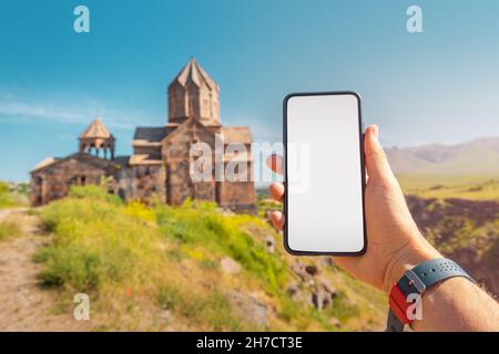 La main du voyageur tient un smartphone avec un écran vierge sur le fond d'une ancienne église arménienne.Concept d'application mobile pour touris Banque D'Images
