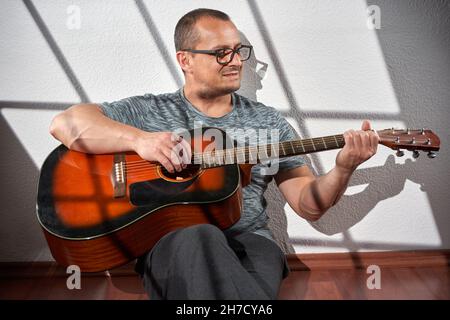 Homme jouant de la guitare acoustique, assis sur le sol, avec la lumière venant de la fenêtre projetée sur le mur Banque D'Images