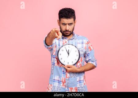 Strict bossy sombre cheveux jeune homme adulte avec la barbe tenant l'horloge murale dans les mains et pointant vers l'appareil photo à vous, gestion du temps, vous êtes en retard.Studio d'intérieur isolé sur fond rose. Banque D'Images