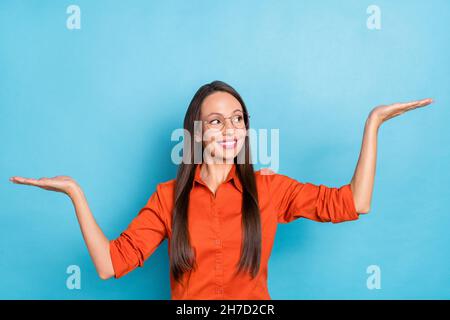 Photo de douce brillante femme mûre vêtue rouge chemise verres montrant les bras balance regardant l'espace vide souriant isolé couleur bleu arrière-plan Banque D'Images