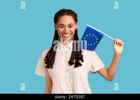 Drapeau de l'Union européenne.Adorable femme souriante satisfaite avec des dreadlocks noirs tenant le drapeau de l'Europe, regardant la caméra, portant une chemise blanche.Studio d'intérieur isolé sur fond bleu. Banque D'Images