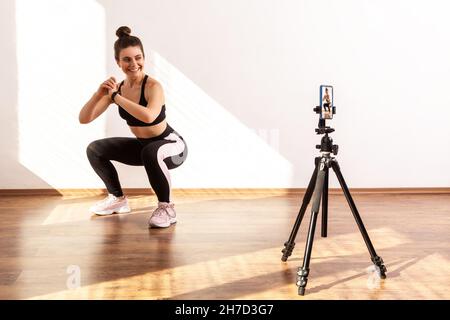 Entraîneur souriant enseignant la technique correcte de squating, enregistrement vidéo tutoriel d'entraînement, port de haut de sport noir et collants.Prise de vue en studio pleine longueur éclairée par la lumière du soleil depuis la fenêtre. Banque D'Images