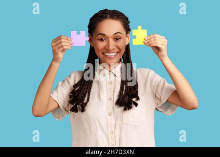 Souriant satisfait femme attrayante avec des dreadlocks noirs montrant à la caméra deux pièces de puzzles, la tâche de résolution, le port de chemise blanche.Studio d'intérieur isolé sur fond bleu. Banque D'Images