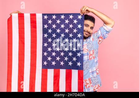 Portrait d'un homme attrayant tenant le drapeau des États-Unis entre les mains, regardant l'appareil photo avec un sourire crasseux, fier de sa sécurité sociale dans le pays, se sentant en sécurité.Studio d'intérieur isolé sur fond rose. Banque D'Images
