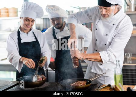 Chef assaisonnement de la viande sur une poêle à frire près de collègues interraciaux souriants avec du romarin dans la cuisine Banque D'Images