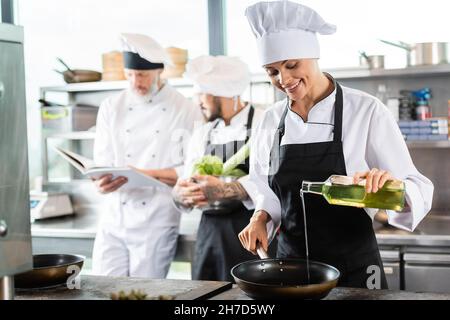 Chef souriant dans un tablier versant de l'huile d'olive sur une poêle près de collègues flous avec livre de cuisine dans la cuisine du restaurant Banque D'Images