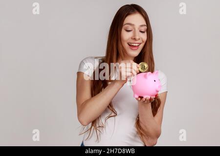 Portrait d'une femme souriante et sombre qui investit dans des bitcoins, mettant la pièce de crypto dorée dans une banque de porc, portant un T-shirt blanc.Prise de vue en studio isolée sur fond gris. Banque D'Images