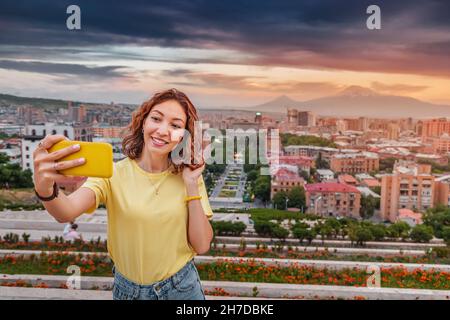 Jeune femme souriante prend une photo de selfie à l'arrière-plan du paysage urbain à Erevan avec une vue sur le célèbre Mont Ararat pendant un soleil coloré Banque D'Images