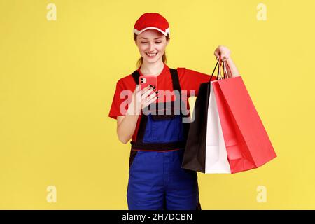 Portrait d'une femme de livraison optimiste posant avec des sacs d'achats et un smartphone dans les mains, en utilisant le téléphone cellulaire vérifiant l'adresse, portant une combinaison et une casquette.Studio d'intérieur isolé sur fond jaune. Banque D'Images