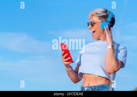 Souriante, jolie jeune femme aux cheveux blonds courts vêtue d'un t-shirt court montrant son bouton du ventre, elle s'amuse tout en écoutant de la musique avec son fil bleu Banque D'Images