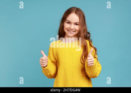 Portrait de charmante petite fille avec des cheveux ondulés montrant les pouces jusqu'à l'appareil photo, exprimant positif, portant jaune style décontracté pull.Studio d'intérieur isolé sur fond bleu. Banque D'Images