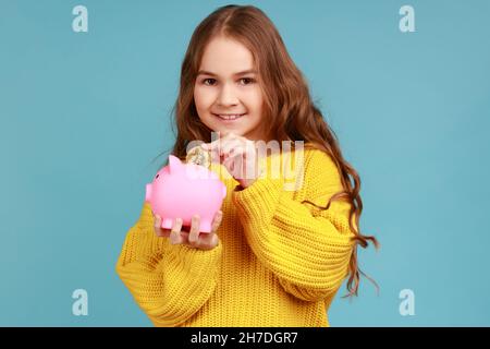 Portrait d'une petite fille mettant la pièce de crypto dorée dans la banque de piggy, regardant l'appareil photo avec le sourire, portant le chandail jaune de style décontracté.Studio d'intérieur isolé sur fond bleu. Banque D'Images