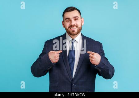 C’est moi.Jeune homme ambitieux avec barbe portant un costume de style officiel avec un sourire éclatant sur le visage pointant des doigts sur lui-même, homme d'affaires confiant.Studio d'intérieur isolé sur fond bleu. Banque D'Images