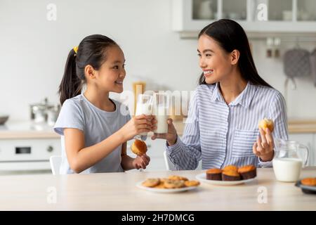 Bonne fille asiatique et sa mère boivent du lait tout en étant assise dans la cuisine, en clinquant des verres et en appréciant la boisson de calcium