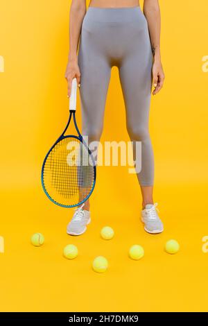 vue partielle de sportswoman dans des leggings gris tenant la raquette tout en se tenant près des balles de tennis sur jaune Banque D'Images