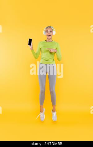 vue intégrale de la sportswoman stupéfait dans les écouteurs pointant vers le téléphone portable avec l'écran de banque sur jaune Banque D'Images