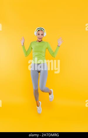vue intégrale de la femme excitée et sportive dans un casque regardant l'appareil photo tout en sautant sur le jaune Banque D'Images